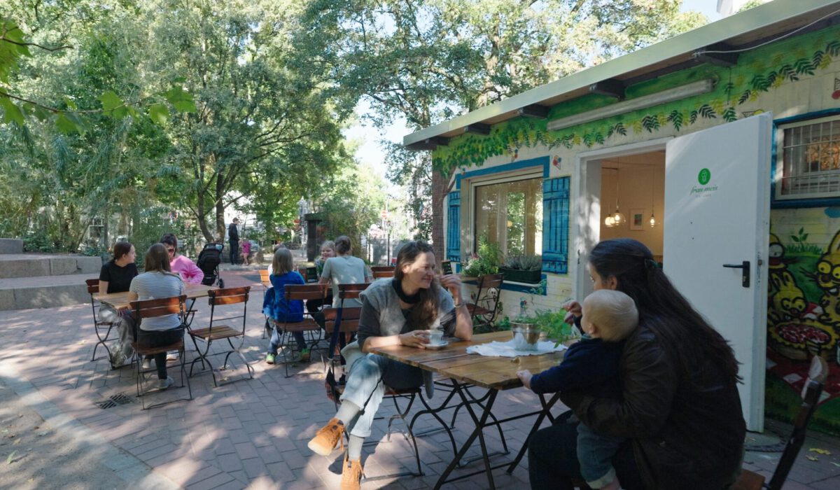 Gäste sitzen draußen zwischen dem Spielpaltz links und Frau Meis Eis · Café rechts an Tischen Stühlen