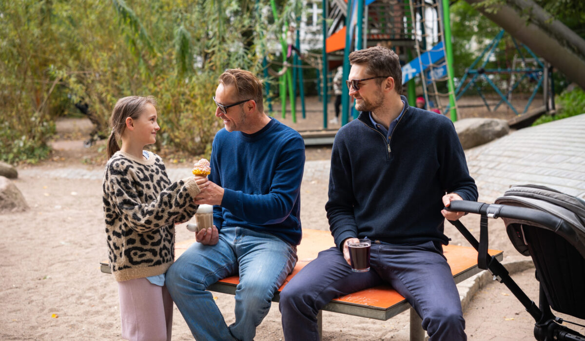 Zwei Männer und ein Kind sitzen vor dem Spielplatz auf einer Bank, das Mädchen reicht ihrem Vater ein Eis, der andere Mann hält einen Kinderwagen fest